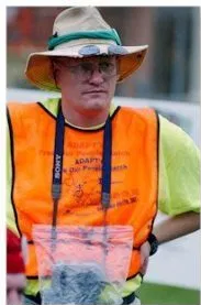 Picture of Tim Wheat A man with a wide brimmed hat, eye glasses, a yellow top and orange vest stands outdoors looking to the distance.