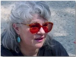 A woman with grey hair pulled back into a ponytail and red lensed glasses looks off to the right.