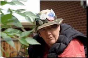 A man with a fisherman's hat covered in buttons, wearing a black vest over a red top. Green leaves are in the foreground.