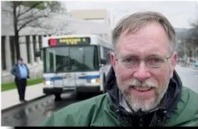 Picture of Jerry Costley A man in a green rain jacket is in the foreground smiling. A city bus is parked in the background.