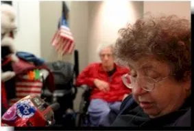 Picture of Bunny McLeod A person in the foreground with short curly hair and eyeglasses looks down at something off camera. A person in a wheelchair wearing a red jacket is in the background with an American flag.