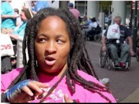 A Black woman with long dark dreadlocs and a pink top is looking to the right mid-sentence