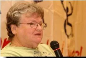 A woman with short blonde hair, eye glasses and a green top speaks into a microphone. The ADAPT logo in the background.