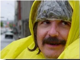 Image of David Wittie A man with long dark hair and a mustache wears a rain cap and yellow rain jacket. He looks off to the right.