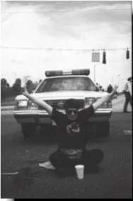 Black and white photo of a protestor sitting on the ground cross-legged, with their arms spread out, in front of a police car