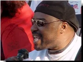 A Black man with a black cap, sunglasses, and white top speaks into a microphone.