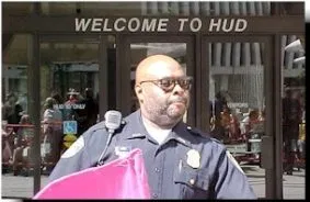 A police officer in uniform stands outside. A "Welcome to HUD" sign is in the background. A part of a pink flag in the foreground.