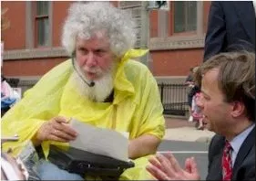 A man with curly grey hair, a white beard, wearing a yellow rain poncho has a microphone headpiece. He speaks to a man in a dark suit kneeling at his side.