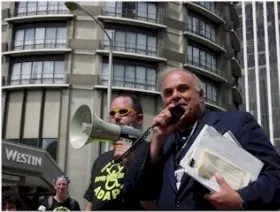 Two men stand in front of a circular building. "Westin" is on the awning. One man holds a stack of papers and speaks into a loud speaker. The other man holds the loud speaker.