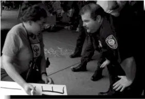 Black and white foto of a person wearing an ADAPT tee shirt holding a handwritten sign. A police officer bends down in front of them.