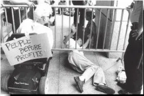 Black and white photo of a person laying on the ground under a barrier gate.  Another person is in a wheelchair in front of the gate with a sign on the back of the wheelchair "People Before Profits"