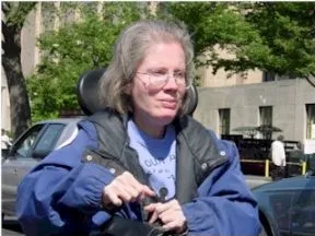 Picture of Diane Coleman of ADAPT A woman with shoulder-length greyish hair and a blue and black jacket sits in a wheelchair outdoors.