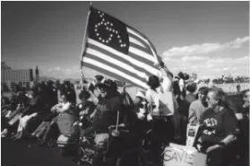 Disabled American flat Black and white photo of protestors in a large crowd. One holds a modified American flag - the stars are in the form of a wheelchair symbol.
