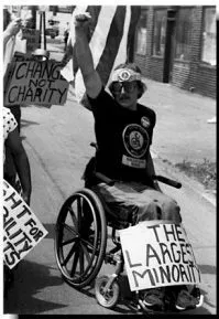 The Largest Minority Black and white photo of a protestor in a wheelchair with their fist raised. There is a sign strapped to their legs saying "The Largest Minority".