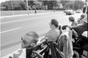 Black and white photo of people in wheelchairs lining the side of a street facing the street.