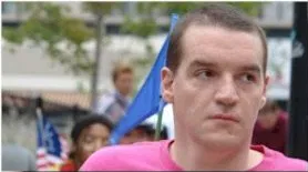 A man with short cropped hair and a pink top looks off into the distance. A blue flag is behind him.