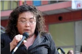A woman with curly hair, glasses, and a dark top speaks into a microphone.
