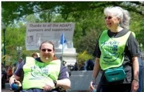 Two people, one in a wheelchair and one standing, wear green ADAPT vests outdoors during a protest
