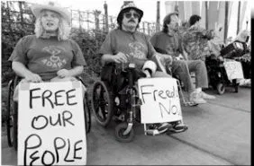 Free Our People. Freedom Now. Black and white picture of protestors in wheelchairs. One woman in an ADAPT tee shirt holds a sign that says "Free Our People". Next to her, a man holds a sign that says "Freedom Now".