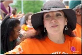 A woman with long dark hair, a hat, and orange top gazes pass the camera. People with similar orange tops are in the background.