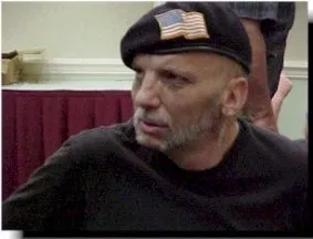 A man in a black beret, with an embroidered American flag, and dark top looks off to the left.