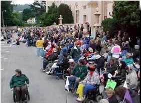 A large group of people in wheelchairs, some standing, outside of a building.