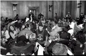Black and white image of a large group of people in a building. Some in wheelchairs. Some not.  A person facing the camera wears a black hat and an ADAPT logo top.