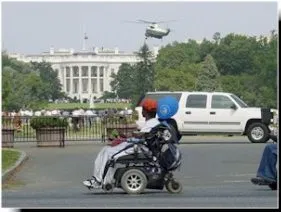 A person in power wheelchair, red cap, white top and light pants stops in the middle of the road.