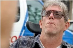 A man with short hair, sunglasses, and a dark top looks up and away from the camera. A blue and white vehicle is in the background.