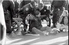 Black and white photo of protestors, wheelchair users, sitting in the middle of a street. One is being lifted by cops up off the ground.