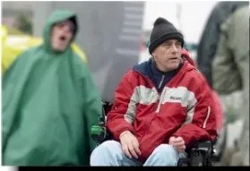 Picture of Randy Alexander A man in a black winter hat and red and white jacket sits in a wheelchair looking off to the right. Another person in a green rain poncho is shouting in the background.