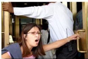 A woman with long dark hair is in a doorway, holding the door open with one hand and shouting. A man in a white top and dark pants is in the background holding on to another door.